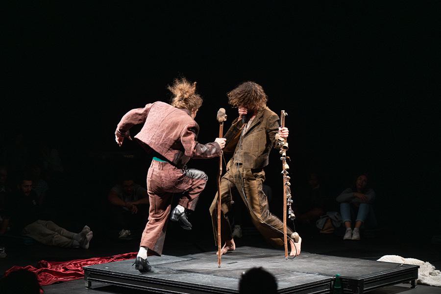 Performance image from Mirlitons featuring Hainaux and Chaignaud performing in the center of a theater on a small square stage. Silhouettes of audience members are seen in the foreground and background. Chaignaud faces away from the camera as he lifts on leg in the air, tap dancing. He wears pink pants and a jacket, holding a large wooden stick. Hainaux faces towards the camera and Chaignaud, wearing a brown suit and holding a wooden stick with bells attached to it. He beatboxes into a microphone.