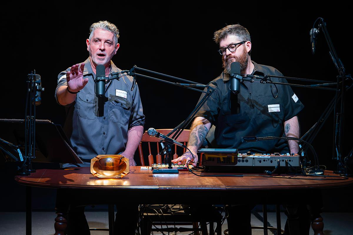 Two male performers sit at a podcast-style table with microphones, sound equipment, and props, delivering a live multimedia performance in front of a colorful, abstract projection